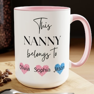 May include: White ceramic mug with a pink rim and handle. The mug has black text that reads "This NANNY belongs to" with three hearts below the text. The hearts are pink, pink, and blue with the names "Olivia", "Sophia", and "Jason" written inside.