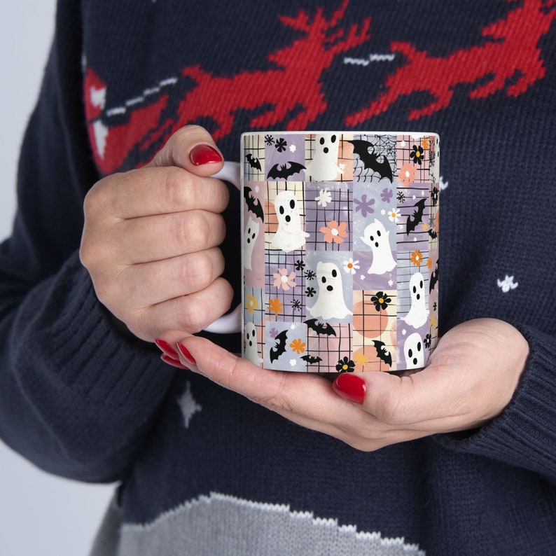 K&ouml;nnte beinhalten: Wei&szlig;e Keramiktasse mit einem Patchwork-Design aus Geistern, Flederm&auml;usen, Blumen und Spinnweben in Pastellfarben. Die Tasse hat einen wei&szlig;en Henkel und wird von einer Person gehalten, die einen marineblauen Pullover tr&auml;gt.