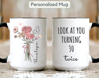 May include: White ceramic mug with black handle and rim. One side features a floral bouquet with pink roses and the name "Penelope". The other side reads "LOOK AT YOU TURNING 30 twice". A personalized gift.