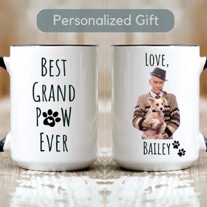 Peut inclure: Deux tasses en céramique blanche avec des poignées noires. La tasse de gauche porte le texte "Best Grand Paw Ever" avec un motif d'empreinte de patte. La tasse de droite porte le texte "Love, Bailey" avec un motif d'empreinte de patte et une photo d'un homme tenant un petit chien.