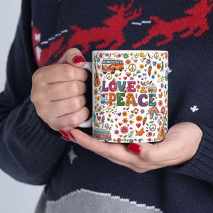May include: White ceramic mug with a colorful "Love & Peace" design. The design includes a peace sign, flowers, musical notes, and a retro van. The mug is held by a person wearing a dark blue sweater.