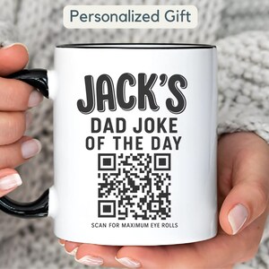 May include: A white ceramic mug with a black handle and rim. The front features the black text 'JACK'S DAD JOKE OF THE DAY' above a square QR code, followed by the text 'SCAN FOR MAXIMUM EYE ROLLS' at the bottom of the design.