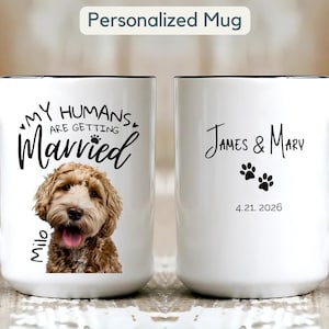 Könnte beinhalten: Weißer Keramikbecher mit schwarzem Griff und Rand. Der Becher zeigt den Text "MY HUMANS ARE GETTING Married" und einen Hund namens Milo. Die andere Seite hat die Namen "James & Mary", Pfotenabdrücke und das Datum 4.21.2026.