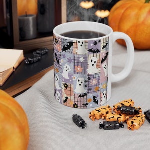 K&ouml;nnte beinhalten: Wei&szlig;e Keramiktasse mit einem Halloween-Motiv. Die Tasse hat ein Patchworkmuster mit Geistern, Flederm&auml;usen und Blumen in Pastellfarben. Die Tasse ist mit einer dunklen Fl&uuml;ssigkeit gef&uuml;llt und steht auf einer hellgrauen Oberfl&auml;che.