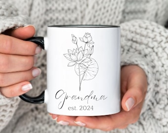 Puede incluir: Taza de cerámica blanca con borde y asa negros. La taza presenta un dibujo de línea negra de una flor de loto y el texto "Grandma est. 2024".