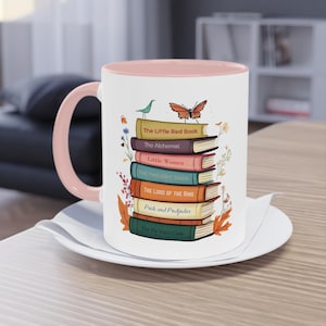 Può includere: Una tazza in ceramica bianca con bordo e manico rosa. La tazza presenta una pila di libri con titoli che includono "The Little Red Book", "The Alchemist", "Little Women", "The Twilight Saga", "The Lord of the Rings", "Pride and Prejudice" e "The Da Vinci Code".
