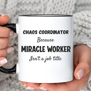Può includere: Tazza in ceramica bianca con manico e bordo neri. La tazza presenta il testo "CHAOS COORDINATOR Because MIRACLE WORKER Isn't a job title" in nero. Un'idea regalo umoristica, tenuta da una persona.