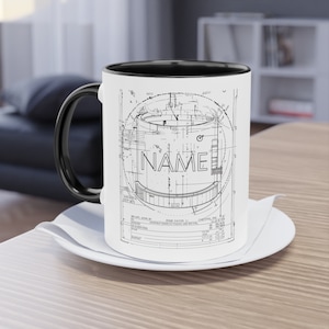 Könnte beinhalten: Weißer Keramikbecher mit schwarzem Henkel und Innenraum, mit einem Design im Blaupausenstil mit dem Wort "NAME". Der Becher steht auf einer weißen Untertasse, ideal für Kaffee oder Tee.