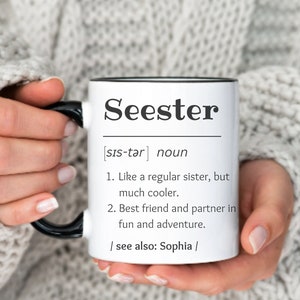 以下が含まれることがあります： 黒いハンドルと縁が付いた白いセラミックマグで、黒い文字で「Seester」という言葉が特徴です。マグには定義が含まれています。「普通の姉妹のように、しかしずっとクール。」と「最高の友達と楽しさと冒険のパートナー。」