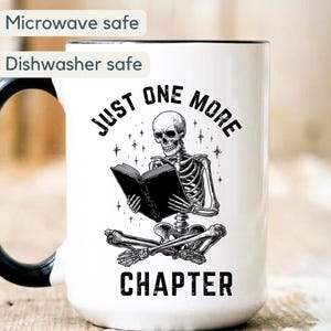 May include: White ceramic mug with a black handle and rim. The mug features a black and white illustration of a skeleton reading a book, with the text "JUST ONE MORE CHAPTER". The mug is microwave and dishwasher safe.