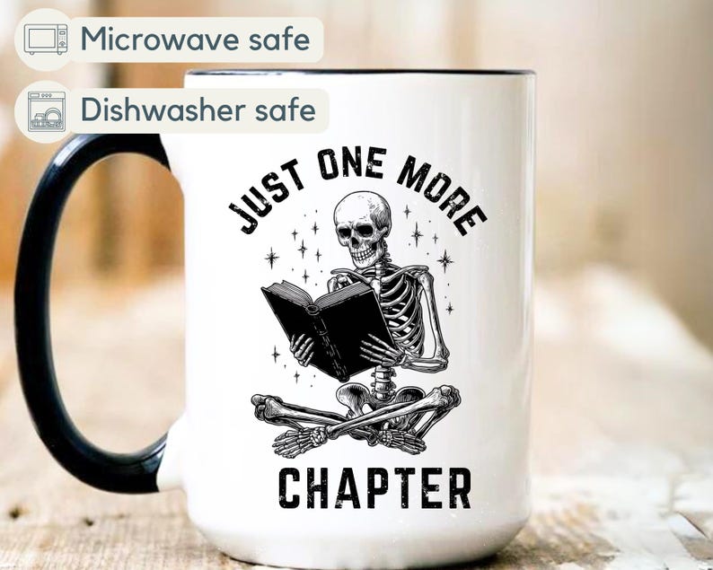 May include: White ceramic mug with a black handle and rim. The mug features a black and white illustration of a skeleton reading a book, with the text "JUST ONE MORE CHAPTER". The mug is microwave and dishwasher safe.