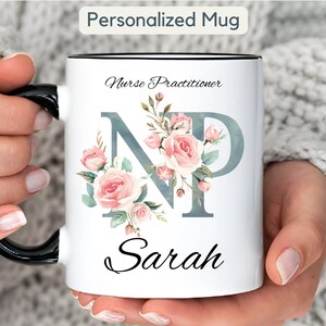 Puede incluir: Taza de cerámica blanca con asa y borde negros, con el texto "Nurse Practitioner" y el nombre "Sarah". La taza tiene un diseño floral con rosas rosas y las letras "NP". Las palabras "Personalized Mug" están en la parte superior.