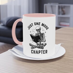 May include: White ceramic mug with a pink interior and handle. The mug features a black illustration of a skeleton reading a book, with the text "JUST ONE MORE CHAPTER" in a circular design.
