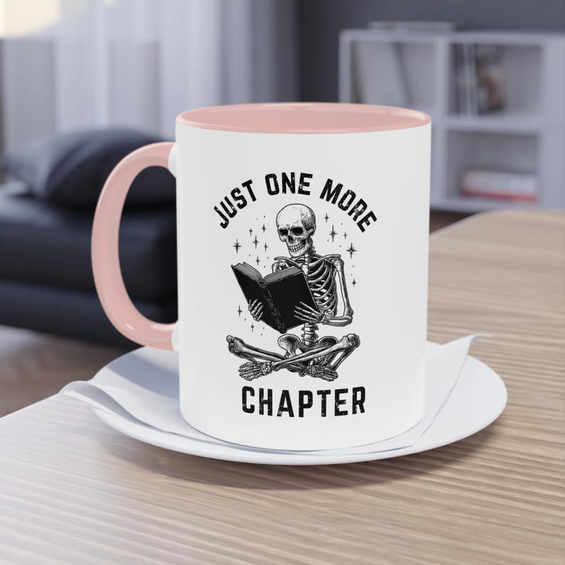 May include: White ceramic mug with a pink interior and handle. The mug features a black illustration of a skeleton reading a book, with the text "JUST ONE MORE CHAPTER" in a circular design.