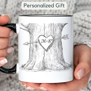 Peut inclure: Mug en céramique blanche avec une anse et un bord noirs. La tasse présente une illustration en noir et blanc d'un tronc d'arbre avec un cœur sculpté, percé d'une flèche. Le cœur contient les initiales "M+S". Le texte "Personalized Gift" est en haut.