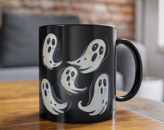 May include: Black ceramic mug featuring a whimsical design of floating cartoon ghosts. The ghosts are white with dark blue outlines and facial features. The mug has a black handle and is sitting on a wooden surface.