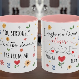 May include: Two white ceramic mugs with pink interiors and handles. One mug reads "You seriously live too damn far from me" with floral and heart accents. The other mug says "I wish we lived closer" with state outlines and names.