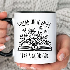 May include: White ceramic mug with a black handle and rim. The mug features the text "SPREAD THOSE PAGES LIKE A GOOD GIRL" in black, along with a floral illustration above an open book. The mug is held by a person.