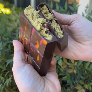 May include: A dark chocolate bar with colourful accents, split open to reveal a pistachio filling. The bar is held in two hands, showcasing the contrasting textures and colours of the treat.