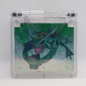 Könnte beinhalten: Ein durchsichtiger Game Boy Advance SP aus Plastik mit einem holografischen Bild eines grün-blauen Pokemon-Charakters auf dem Bildschirm. Der Charakter ist von weißen Sternen auf grünem Hintergrund umgeben.