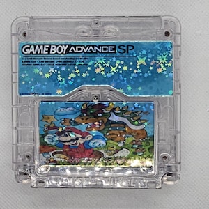 Puede incluir: Un cartucho de Game Boy Advance SP en una carcasa de plástico transparente. La etiqueta superior es azul con el texto "GAME BOY ADVANCE SP" y estrellas holográficas. La etiqueta inferior presenta una colorida ilustración de personajes de videojuegos.