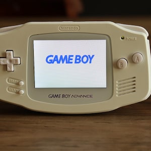 May include: A white Nintendo Game Boy Advance handheld gaming console with a backlit screen displaying the blue text GAME BOY. The device features a directional pad, A and B buttons, Start and Select buttons, and a power indicator light.
