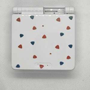 May include: White handheld games console with a flip-up screen. The console is decorated with red, blue, and green triangle-shaped designs. The top has a hinge and a small slot. The bottom has a power button and a small speaker.