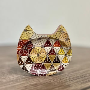 Pode incluir: Um prato de madeira em forma de gato com um padrão geométrico em tons de vermelho, dourado e marrom. O padrão é composto por triângulos e estrelas.