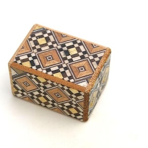 Puede incluir: Una pequeña caja de madera con un patrón geométrico en tonos marrones, negros y blancos. La caja está hecha de madera de marquetería y tiene un borde decorativo.