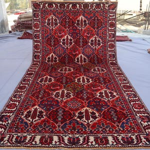 May include: A large, rectangular rug with an intricate, repeating pattern. The rug features a deep red background with accents of blue, cream, and brown. The design includes floral and geometric motifs, creating a rich, detailed texture. The rug is displayed outdoors.