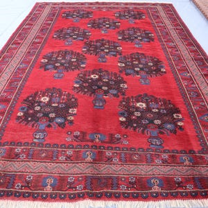 May include: A rectangular red rug with a floral design. The rug features a repeating pattern of stylized floral arrangements in vases, with a border of geometric and floral motifs. The rug has a fringe along the bottom edge.