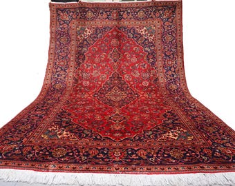 Puede incluir: Una gran alfombra persa rectangular con un diseño de medallón central. La alfombra presenta un campo rojo vibrante con intrincados patrones florales en azul marino, crema y verde. El borde es azul marino oscuro con motivos florales. Flecos blancos.