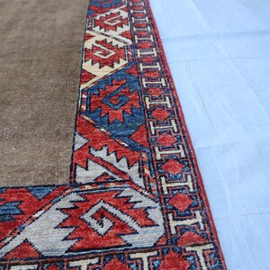 May include: A section of a brown rug with a detailed border. The border features geometric patterns in red, blue, and cream colors. The rug has a fringe along the edge. The rug is likely made of wool or a similar material.