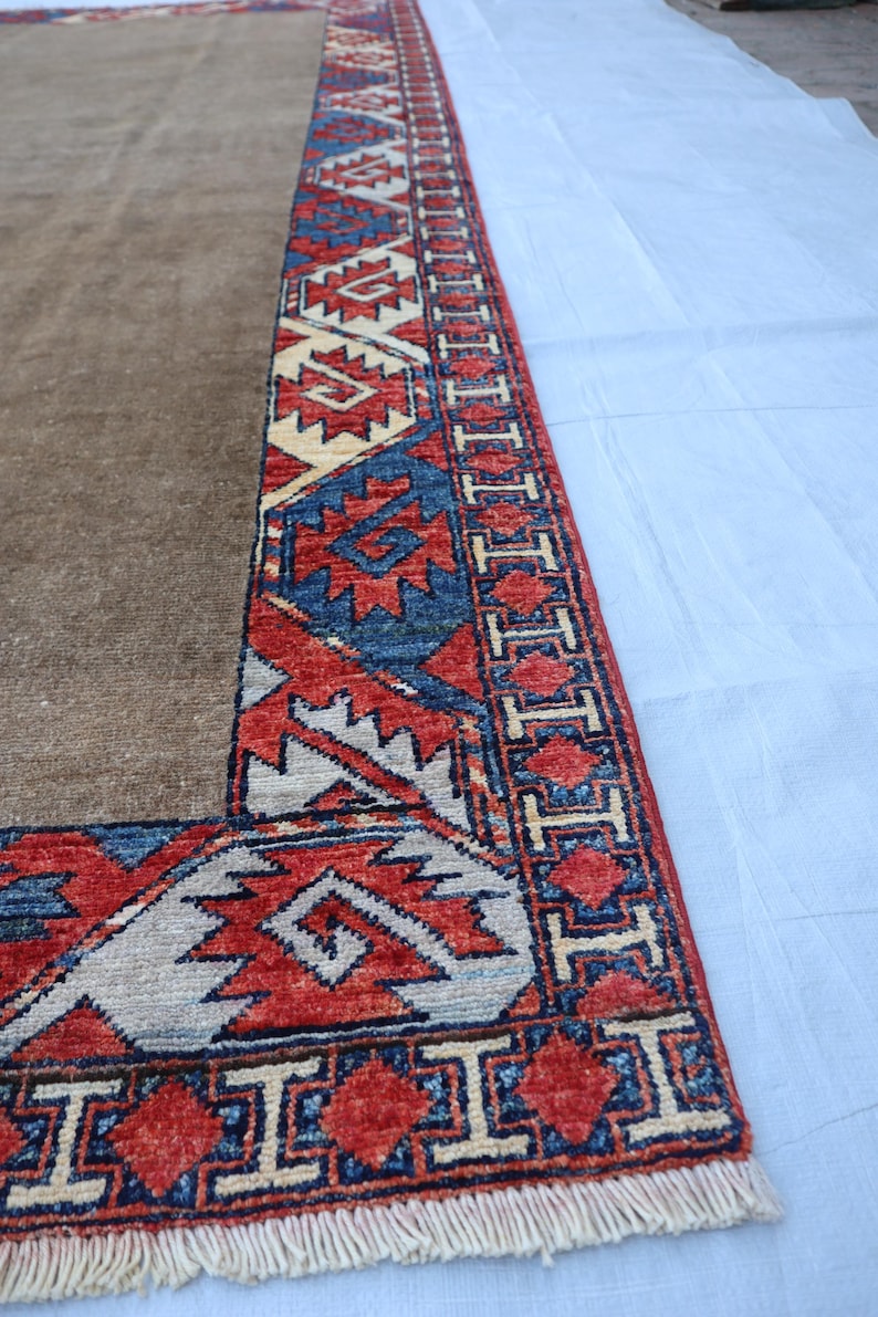 May include: A section of a brown rug with a detailed border. The border features geometric patterns in red, blue, and cream colors. The rug has a fringe along the edge. The rug is likely made of wool or a similar material.