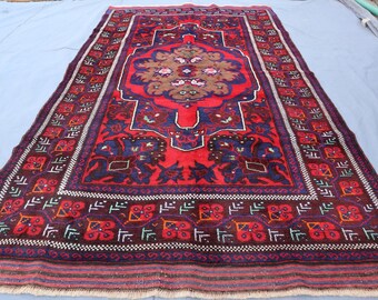Puede incluir: Una alfombra rectangular tejida a mano con un diseño de medallón central. La alfombra presenta un campo rojo vibrante con intrincados patrones en azul, marrón y blanco. El borde es de color púrpura oscuro con motivos geométricos repetidos. La alfombra es probablemente de lana.