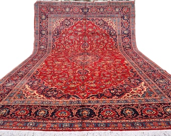 May include: A large, rectangular red rug with an intricate floral pattern. The rug features a central medallion design and a dark blue border with floral motifs. The rug has a cream-coloured fringe along the bottom edge.