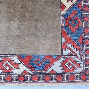 May include: A section of a brown rug with a detailed border. The border features a repeating geometric pattern in red, blue, and cream colors. The rug has a fringe along the bottom edge.