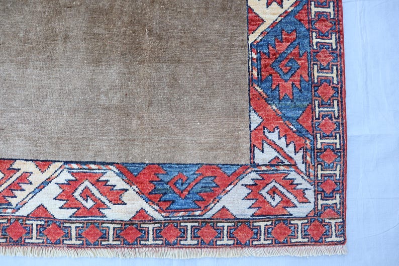 May include: A section of a brown rug with a detailed border. The border features a repeating geometric pattern in red, blue, and cream colors. The rug has a fringe along the bottom edge.