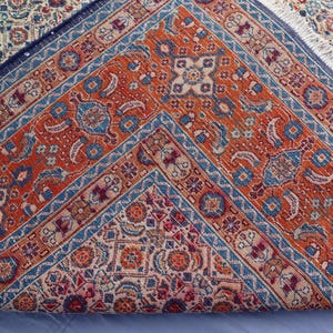 May include: A woven area rug folded to reveal a vibrant orange central field with intricate blue, cream, and red geometric patterns. The rug features a dark blue border with repeating motifs and a white fringe along the folded edge.