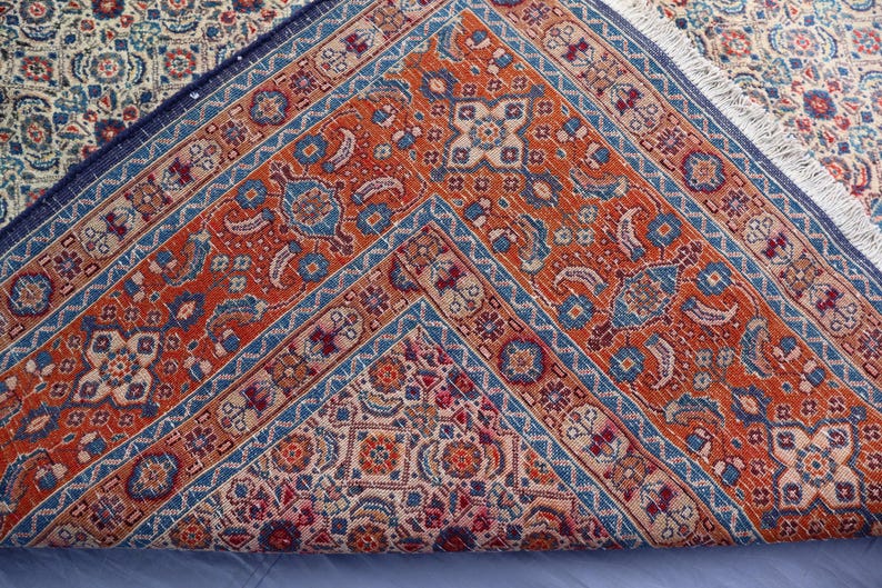May include: A woven area rug folded to reveal a vibrant orange central field with intricate blue, cream, and red geometric patterns. The rug features a dark blue border with repeating motifs and a white fringe along the folded edge.