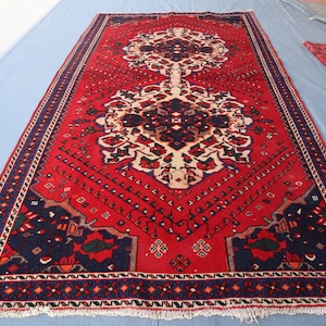 May include: A red, blue and white patterned rug with a traditional design. The rug has a fringed border and is made of wool.