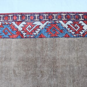 May include: A section of a brown rug with a detailed border. The border features a repeating pattern of red, white, and blue geometric shapes. The rug has a neutral color palette.