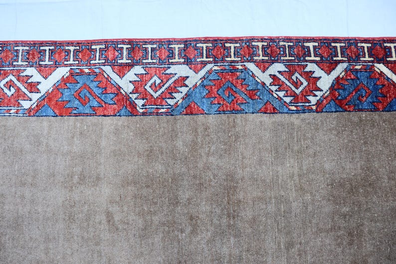May include: A section of a brown rug with a detailed border. The border features a repeating pattern of red, white, and blue geometric shapes. The rug has a neutral color palette.