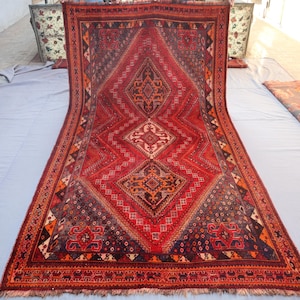 May include: A rectangular, hand-woven rug with a central diamond pattern in shades of red, orange, and navy blue. The rug features intricate geometric designs and a fringed edge. The rug is displayed on a light grey surface.