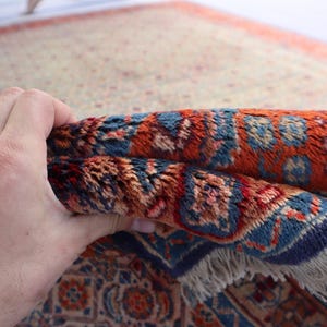 May include: A rectangular area rug featuring a dense pile with a complex pattern in shades of rust orange, deep blue, and cream. The rug has a visible fringe along the edge and a thick, textured weave that shows intricate floral and geometric motifs.