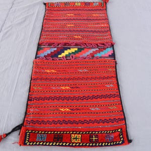 May include: A rectangular, hand-woven textile with a vibrant red background. The piece features intricate geometric patterns in various colors, including blue, yellow, and green. The edges are finished with a black border and tassels, showcasing traditional craftsmanship.