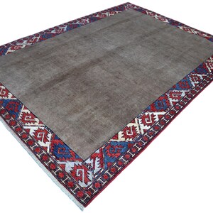 May include: A rectangular rug with a central field of muted brown. The border features a repeating geometric pattern in red, white, and blue. The rug is likely made of woven material and is suitable for a living room or bedroom.