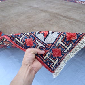 May include: A beige rectangular rug with a detailed border in red, blue, and white geometric patterns. The rug's corner is lifted, revealing the intricate design and fringe.