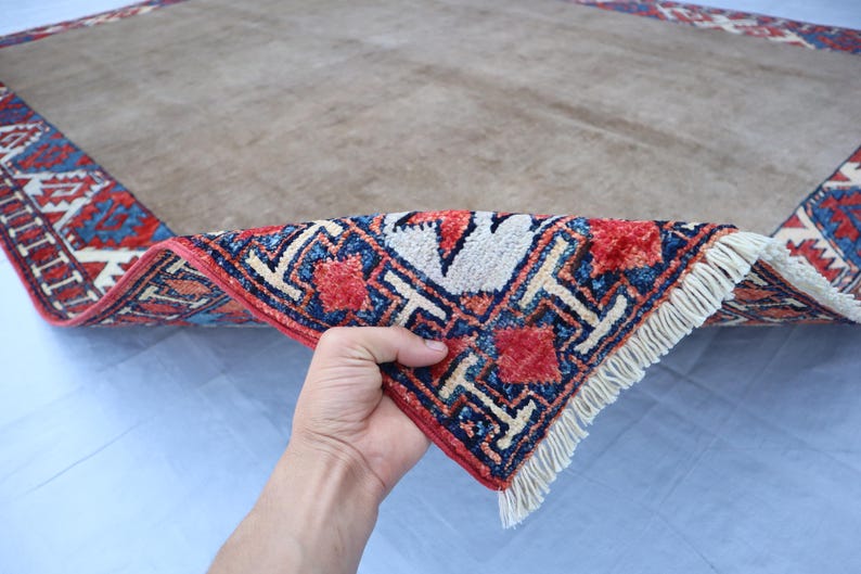 May include: A beige rectangular rug with a detailed border in red, blue, and white geometric patterns. The rug's corner is lifted, revealing the intricate design and fringe.