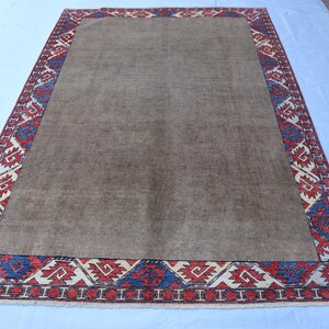 May include: A rectangular, brown rug with a patterned border. The border features a repeating geometric design in red, blue, and white. The rug is likely made of woven material and is suitable for a living room or bedroom.